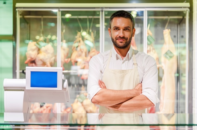 Foto macellaio sicuro. giovane macellaio sorridente che tiene le braccia incrociate e guarda la telecamera mentre si trova alla cassa del supermercato con carne sullo sfondo meat