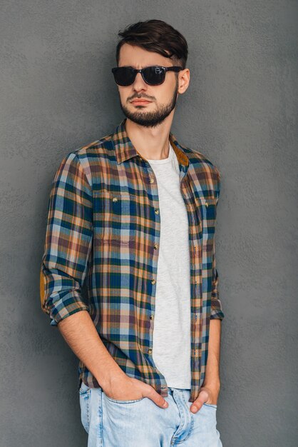 Confident and casual. Confident young man in sunglasses holding hands in pockets and looking away while standing against grey background