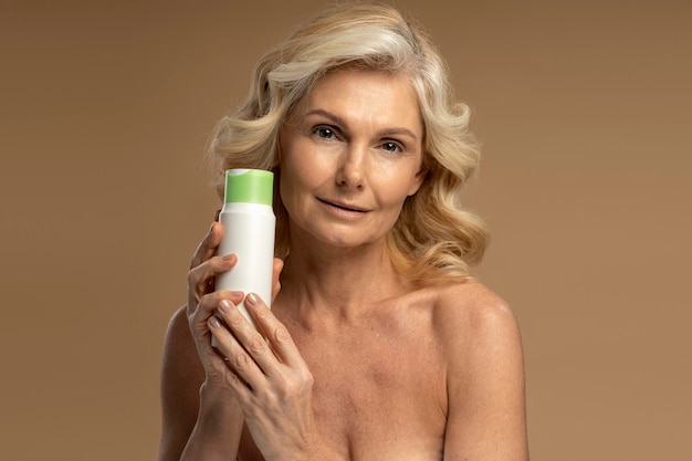 Confident Caucasian 50s woman holding jar of antiaging cosmetic product isolated beige background