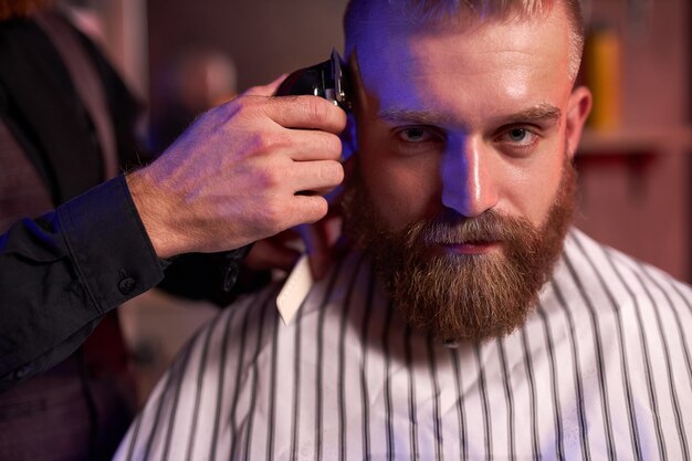 Confident caucasian barber mster cuts hair and beard of men in the barbershop