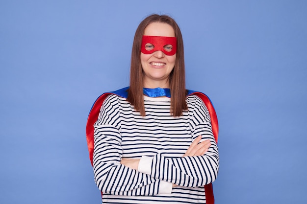 Confident caucasian brown haired woman wearing superhero costume and striped shirt isolated over blue background standing with folded hands looking at camera with happy space
