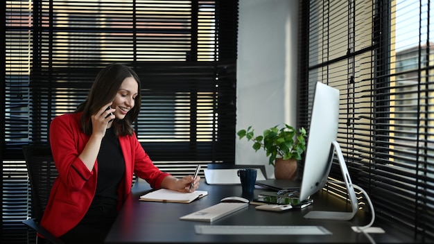 Confident caucasian businesswoman in elegant red suit having pleasant conversation sitting at desk in front of computer