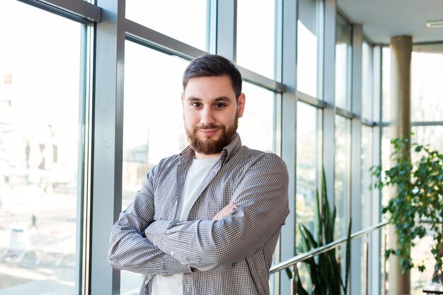 Confident caucasian man standing near full length window in office with plants. Successful bearded young man crossed arms. Handsome businessman smart man in casual shirt.