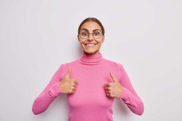 Confident cheerful woman with dark hair smiles pleasanty keeps thumbs up does positive gesture happy for success says very good praises work approves and agrees likes something poses indoor