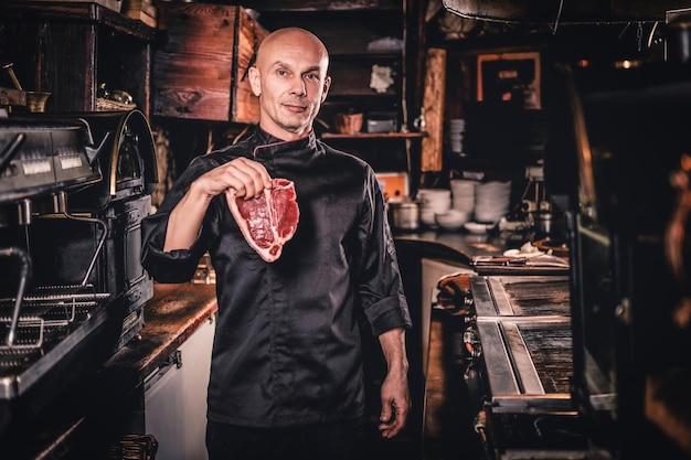 Confident chef wearing uniform holding a fresh piece of meat and looking at a camera in a restaurant kitchen.