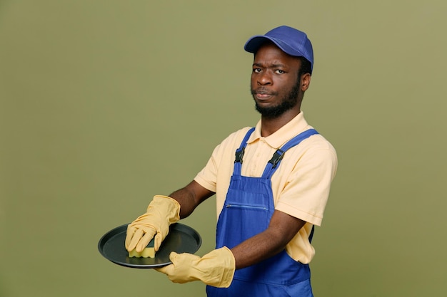 confident cleaning tray with sponge holding sponge on tray young africanamerican cleaner male in uniform with gloves isolated on green background