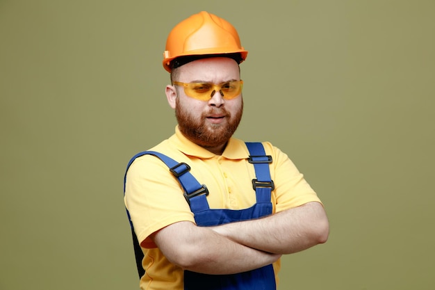 Confident crossing hands young builder man in uniform isolated on green background