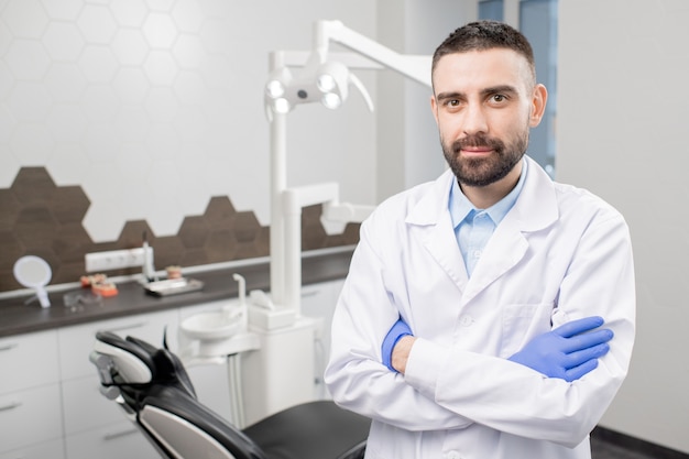 Dentista fiducioso con la barba che incrocia le braccia sul petto mentre è in piedi sul posto di lavoro