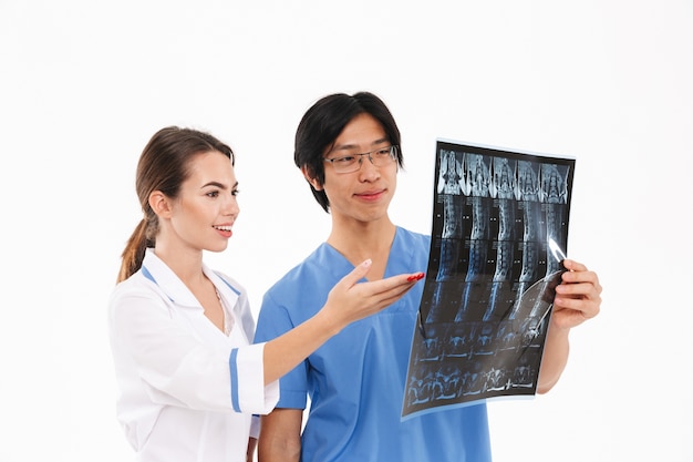 Confident doctors couple wearing uniform standing isolated over white wall, examining x-ray photograph