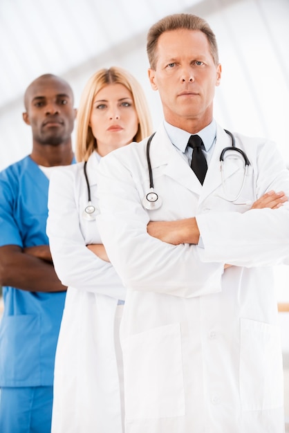 Photo confident doctors team. confident male doctor looking at camera and keeping arms crossed while his colleagues standing in a row behind him