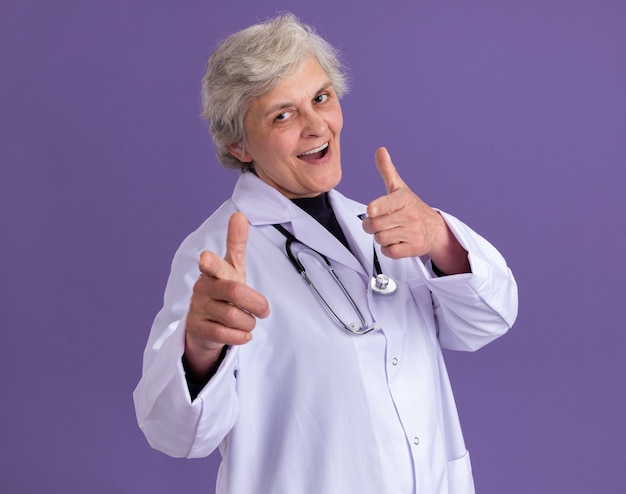 confident elderly woman in doctor uniform with stethoscope pointing at front with two hands isolated on purple wall