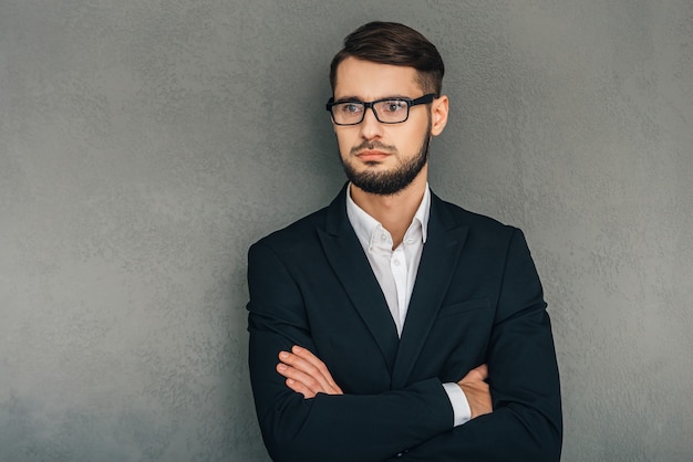 Confident and elegant. Confident young man in glasses looking away and keeping arms crossed while standing against grey background