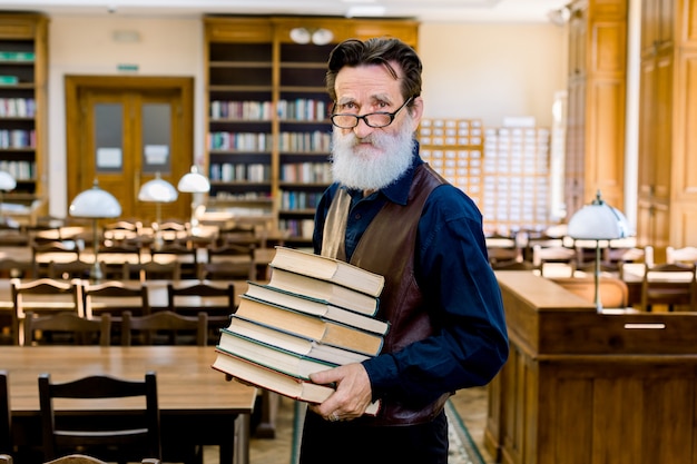 Photo confident elegant librarian of university professor teacher man, wearing stylish clothes, happy to share knowledge, holding stack of different books, standing in vintage library indoors.