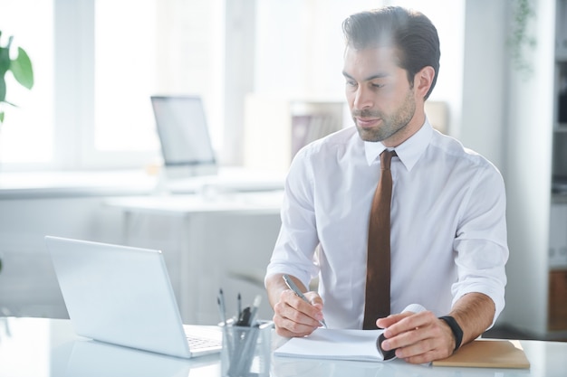 Confident employee making notes while watching online video in front of laptop in office