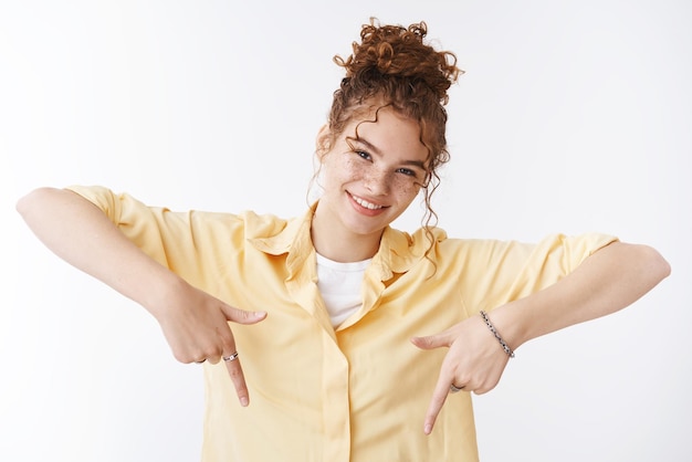 Photo confident energetic positive attractive ginger girl messy curly bun freckles wearing yellow shirt pointing down smiling selfassured offering awesome promo show you perfect advertisement place