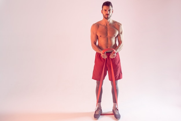 Confident european sportsman doing exercise with resistance band. Young beautiful bearded man with naked sportive torso. Isolated on beige background. Studio shoot. Copy space