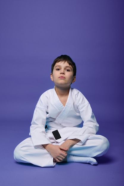 Confident European teenager aikido fighter in white kimono sitting in lotus pose while practicing oriental martial arts on purple background with copy space