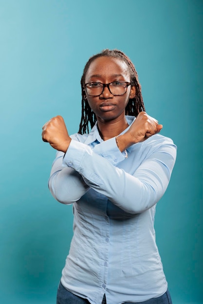 Confident and expressive woman making decline crossed arms symbol while standing on blue background. Serious and confident young adult person showing restriction hands gesture while looking at camera.