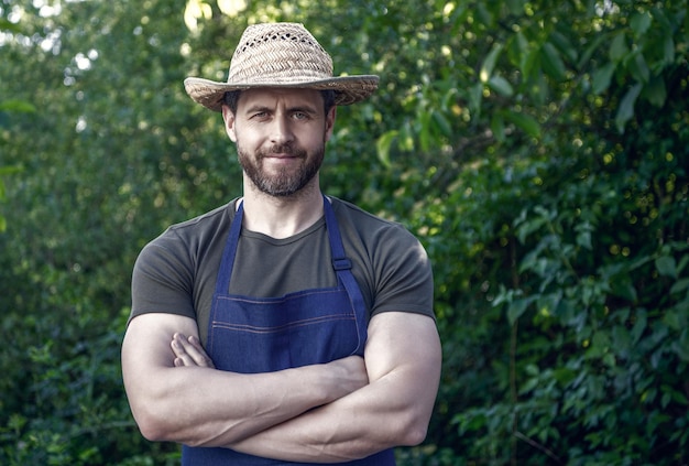 Confident farmer man in farmers hat and apron keeping arms crossed in garden outdoors