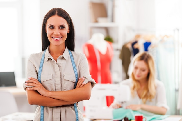 Confident fashion expert. Cheerful young woman keeping arms crossed and smiling while another woman sewing in the background