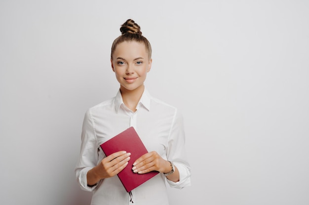 Confident female accountant in white shirt hair in bun holds red notebook with company logs and transactions looks at camera isolated on grey background