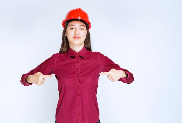 Confident female architect in red hard helmet standing and pointing. 