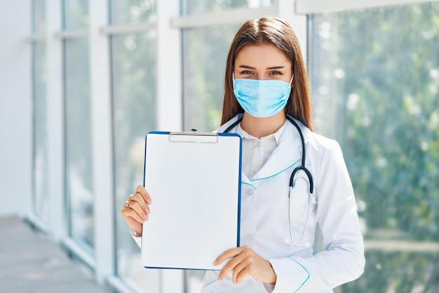 Confident female doctor wearing medical mask showing clipboard\
with diagnosis in hospital background