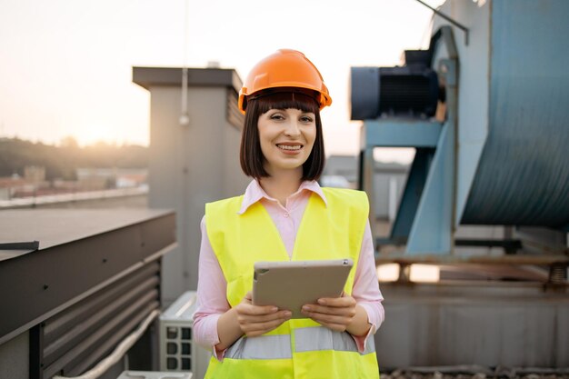 Photo confident female factory worker looking at camera while standing on rooftop ambitious caucasian woman holding personal tablet and checking serviceability of industrial equipment outdoors