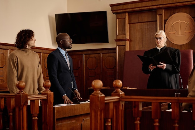 Confident female judge in black mantle standing in front of young intercultural attorney