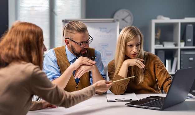 Fiduciosi partner commerciali femminili e maschili che utilizzano laptop moderno durante il briefing in ufficio tre colleghi che condividono idee e discutono della strategia del progetto