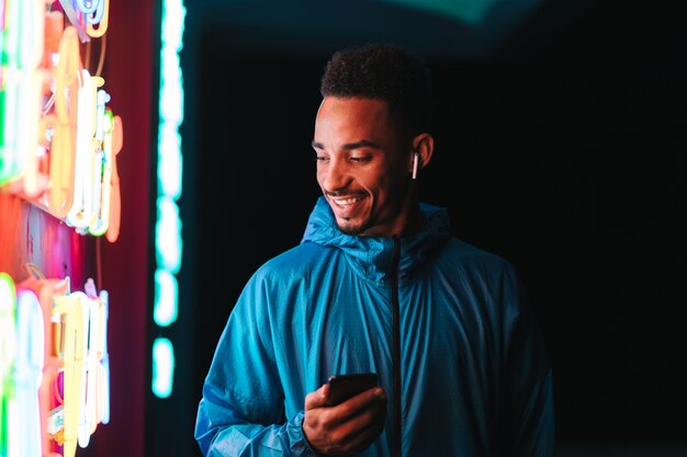 Confident fit young african sports man wearing windbreaker exercising outdoors at night time, using mobile phone