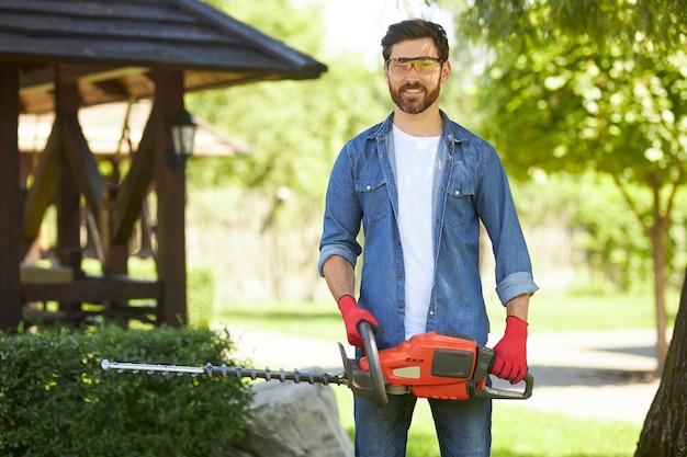 Confident gardener in protective glasses smiling while posing with electric hedge trimmer outdoors