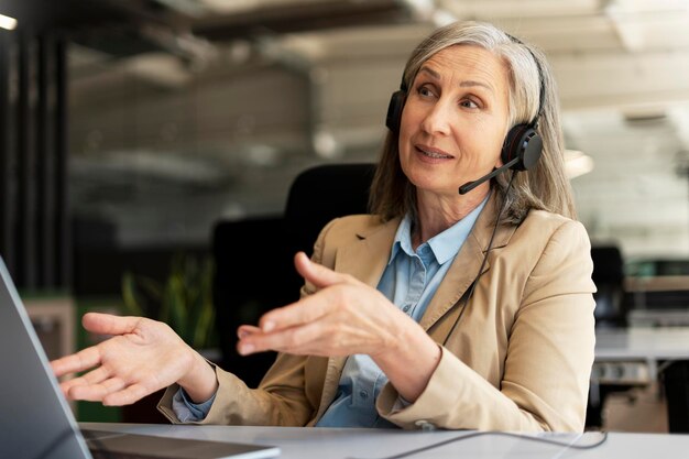 Photo confident gray haired senior businesswoman ceo wearing headphones talking to clients investors or customers via online video conference sitting at desk with laptop in office modern interior