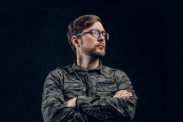 A confident hacker in a stylish military shirt posing with crossed hands and looking sideways. Studio photo with dark background