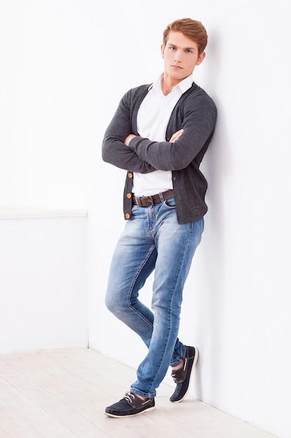 Confident handsome. Full length of handsome young man keeping arms crossed and looking at camera while leaning at the wall