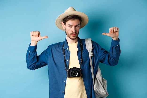 Photo confident handsome guy in summer hat, pointing at himself with bragging look, going to travel on holidays, holding backpack and camera, blue background.