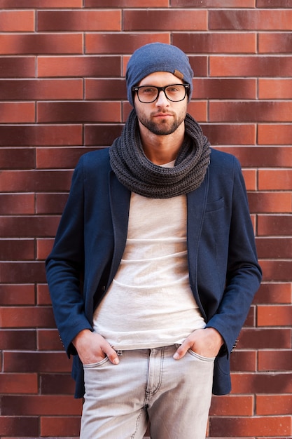 Confident handsome. Handsome young man in smart casual wear holding hands in pockets and looking at camera while standing against brick wall