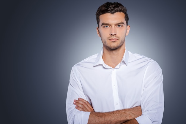 Confident handsome. Handsome young man in white shirt looking at camera and keeping arms crossed while standing against grey background