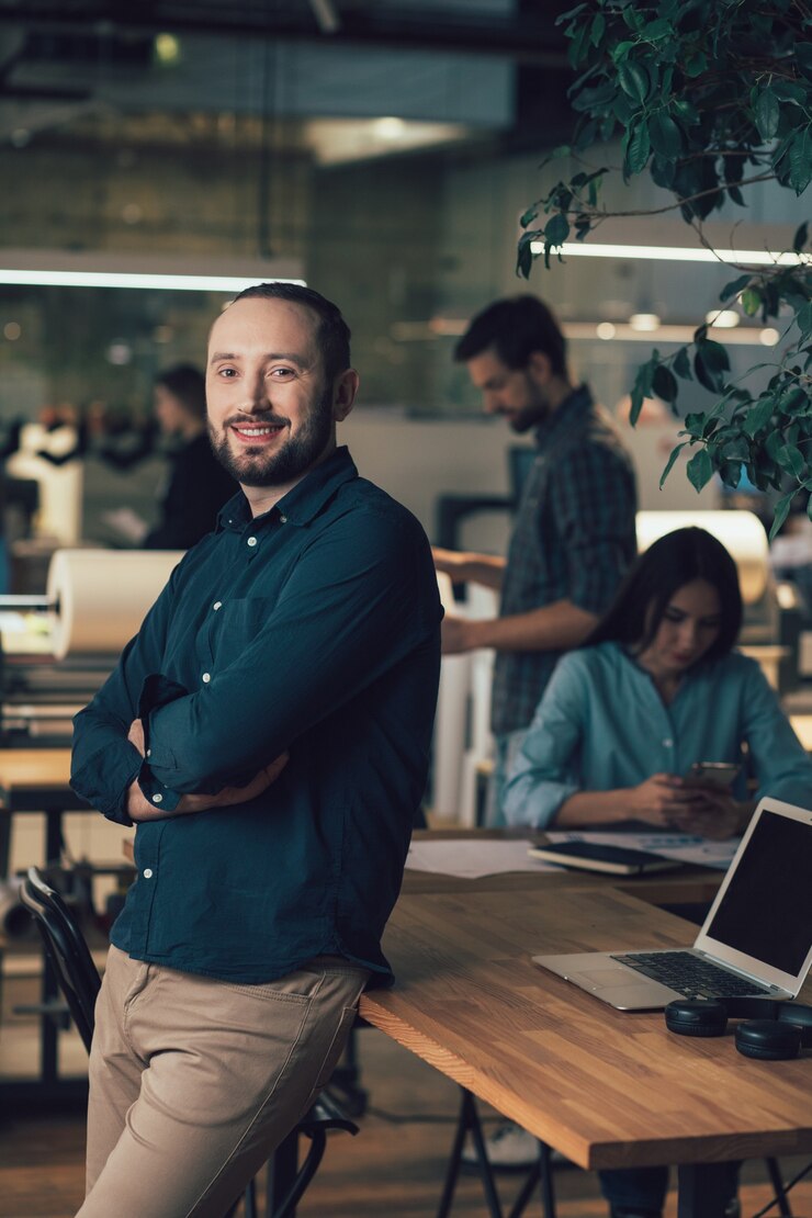 Photo confident handsome man standing by the table in the office and smiling. man and woman on the background busy while working