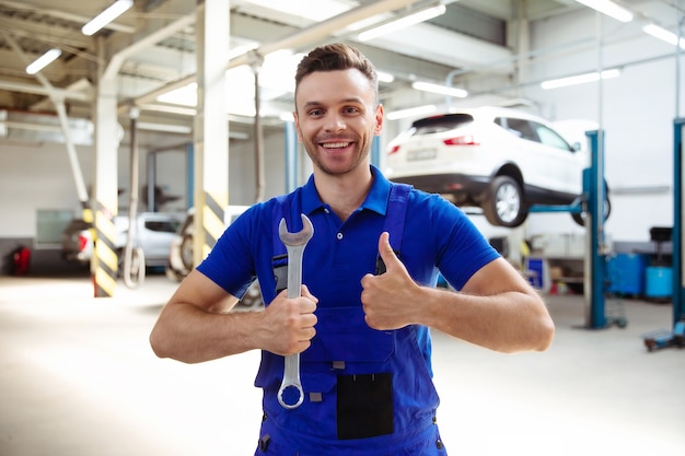 Fiducioso bel giovane ed esperto riparatore di auto in tute da lavoro in posa sullo sfondo di auto sollevate in un servizio di auto