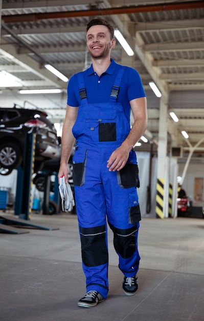 Confident handsome young and experienced car repair worker in work overalls posing against the background of lifted cars in a car service