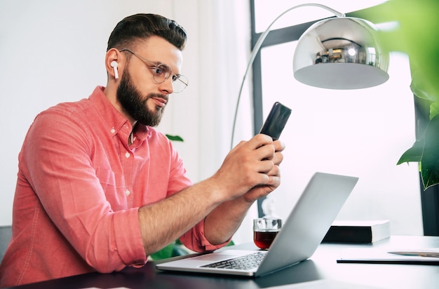 Foto uomo alla moda barbuto fiducioso e felice in occhiali da vista e abbigliamento casual intelligente sta usando il suo smartphone.