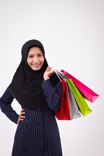 Confident happy smiling muslim woman shopping; studio portrait of islamic woman hand holding shopping bag; happy asian muslim girl doing recreation shopping hobby; asian girl adult woman model