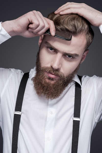Confident in his perfect style. Confident young bearded man combing his hair and looking at camera while standing against grey background
