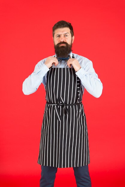 Confident in his recipe. modern cafe concept. cooking modern\
meals. man with beard cook hipster apron. hipster chef cook red\
background. bearded man chef cooking. hipster cooking home or\
restaurant.