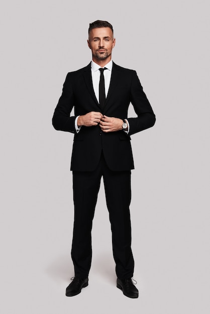 Confident in his style. Full length of handsome young man in full suit buttoning his jacket and looking at camera while standing against grey background