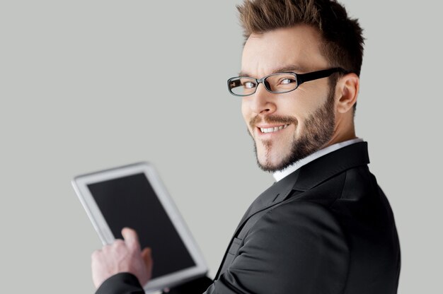 Photo confident in his tablet. rear view of cheerful young man in formalwear and glasses working on digital tablet and looking over shoulder while standing against grey background