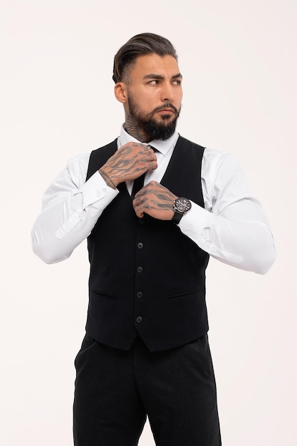 Confident Hispanic bearded man with tattooed hands in white shirt and black trousers adjusting tie