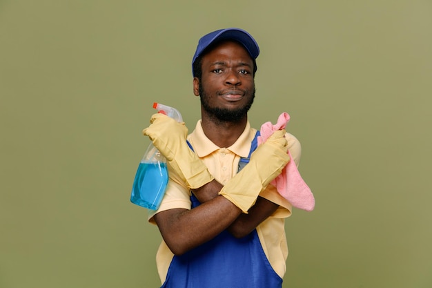 Confident holding cleaning agent with rag young africanamerican cleaner male in uniform with gloves isolated on green background