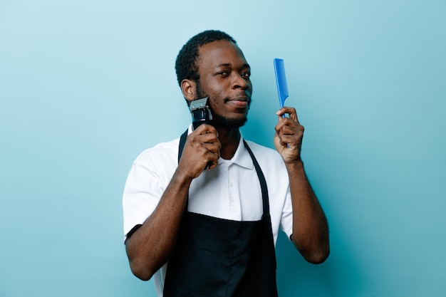 Confident holding comb trimming beard with hair clippers young african american barber in uniform isolated on blue background